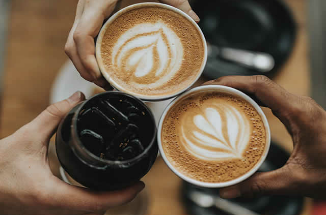 Leute befinden sich am Tisch und halten jeweils Ihre Tasse Kaffee in die Höhe, darunter sind 2 Cappucinos und ein Americano mit Eis.